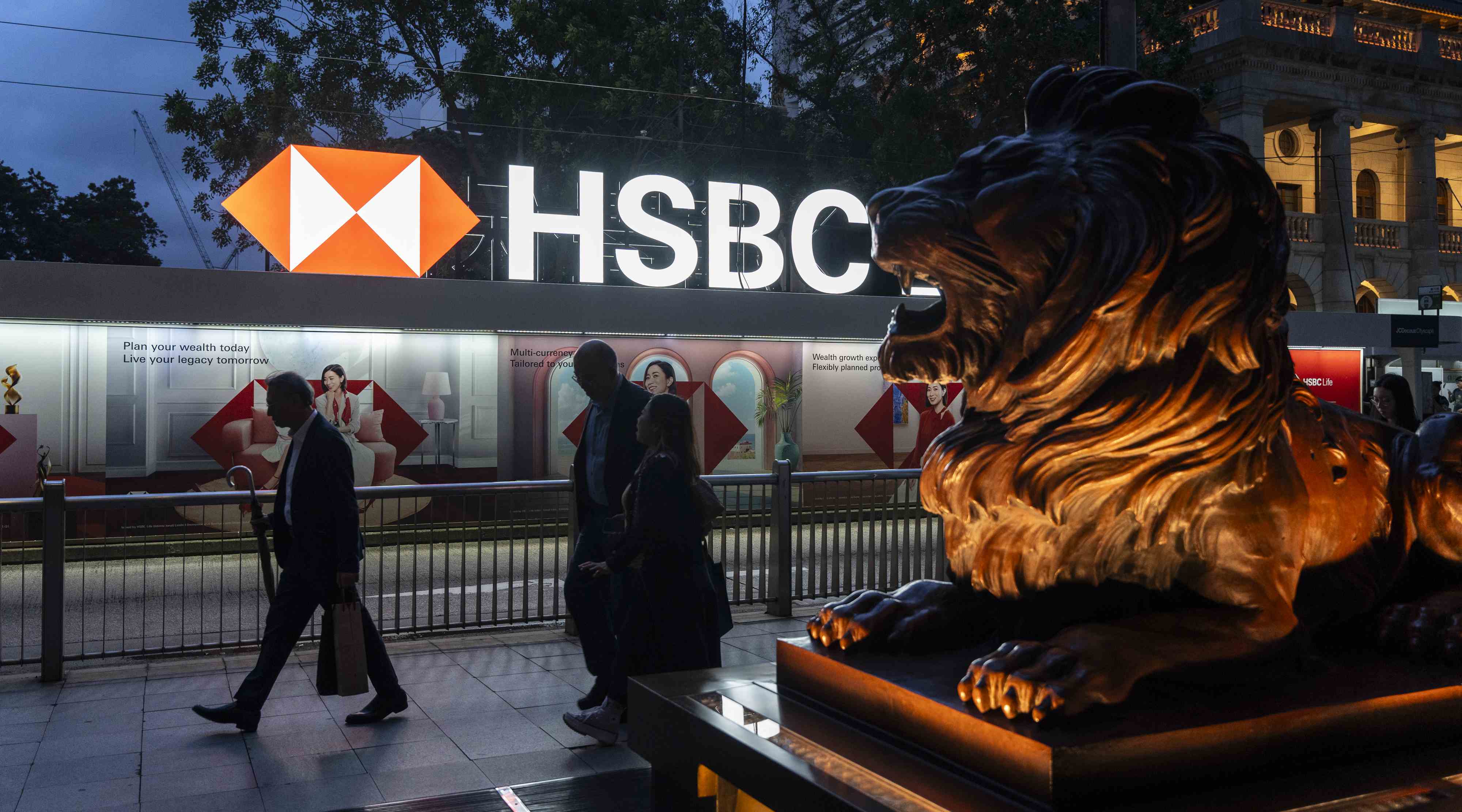 A brightly illuminated HSBC sign is displayed outside a building in Hong Kong, with a lion statue in the foreground and pedestrians walking past during dusk.